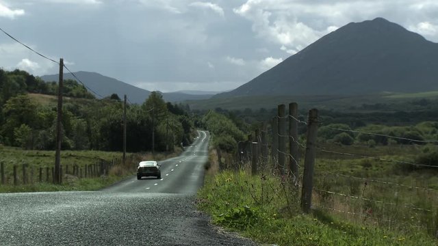 Steady, Wide Shot Of A Car Driving Away From The Camera And Towards A Mountain.