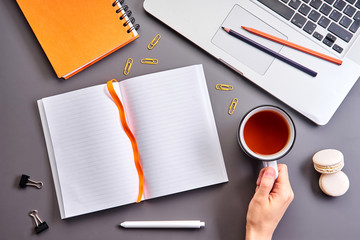 Female hand holding tea cup. Laptop, orange notebook, opened empty notebook and stationery on gray table at home office or workplace. Top view with copy space