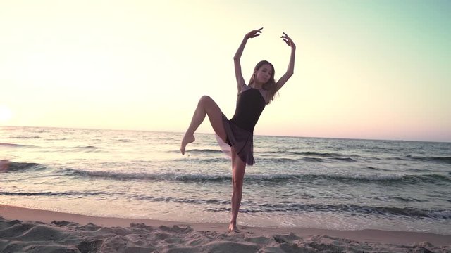 Young pretty ballerina in black dress dancing ballet on sea or ocean sandy beach in morning light. Concept of stretching, art, nature beauty