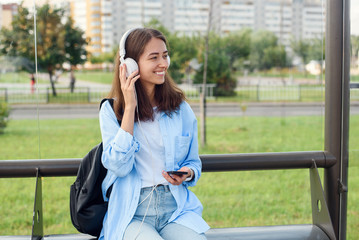 Teen girl listens to the music by white headphones in a public transport station while she waiting for tram.