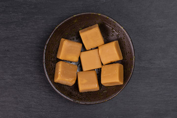 Group of seven whole sweet golden caramel candy in dark ceramic bowl flatlay on grey stone