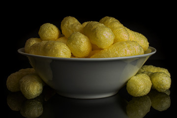 Lot of whole salted yellow corn puff in white ceramic bowl isolated on black glass