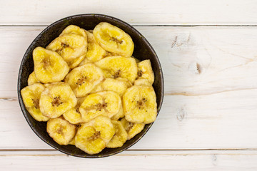 Lot of slices of sweet yellow dry banana in dark ceramic bowl flatlay on white wood
