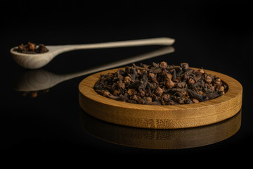 Lot of whole dry brown clove on bamboo coaster in a wooden spoon isolated on black glass