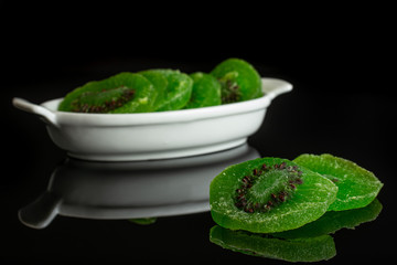 Group of six slices of sweet green candied kiwifruit in white oval ceramic bowl isolated on black glass