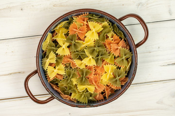 Lot of whole red, yellow and green uncooked farfalle in old pot flatlay on white wood