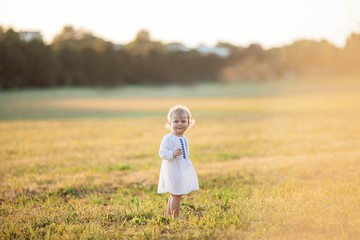 Little curly, blue-eyed girl in a white dress walks in the field. Portrait of a little girl in the sun. Sunset.