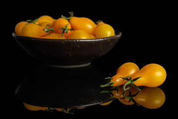 Lot of whole fresh yellow pear tomato two are aside and the rest is in dark ceramic bowl isolated on black glass