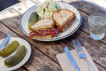 Pastrami sandwich sliced in half on rye bread with mustard on a white plate on a wood table as seen from above. Coleslaw and dill pickle on side. Toothpicks in both halves of sandwich. 