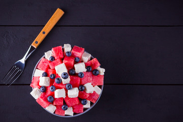 Watermelon salad with feta cheese in gray bowl. Healthy salad with blueberries, watermelon and soft cheese. Summer salad on dark background. Dark and moody