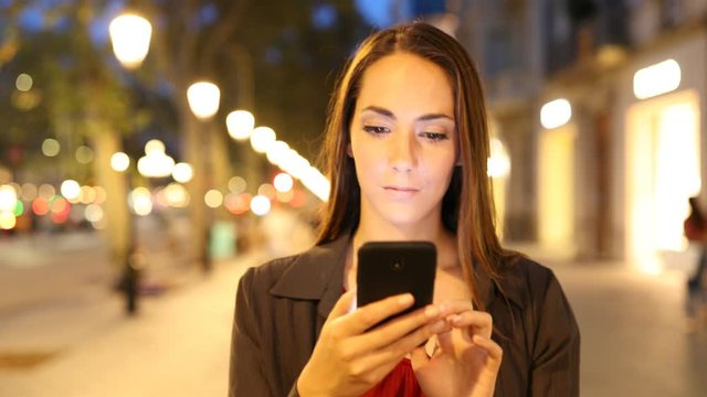 Front view portrait of a woman walking in the night using smart phone in the street
