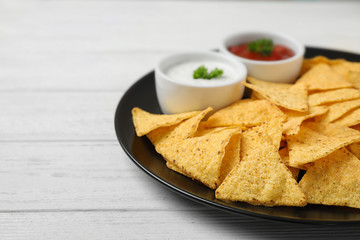 Black plate with tasty Mexican nachos chips and sauces on white wooden table, closeup. Space for text