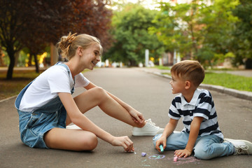 Teen nanny and cute little boy drawing with chalks on asphalt