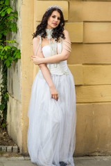 Portrait of Sexy Caucasian Brunette Bride with Tiara Posing Against Old Building. Wearing Beads Necklace and Decorations.