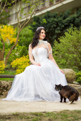 Funny Portrait of Caucasian Bride With Diadem Sitting In Flowers Garden with Kitten Outdoors. Wearing Beads Necklace.