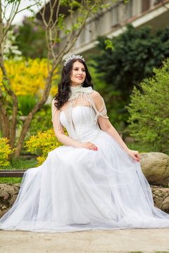 Portrait of Sensual Caucasian Bride With Diadem Sitting In Flowers Garden Outdoors. Wearing Beads Necklace.