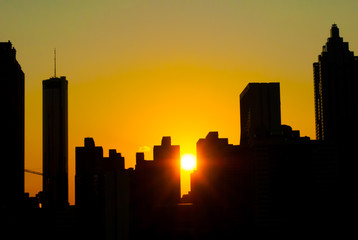 The sun sets on a city skyline which creates a stunning silhouette effect underneath a colorful sky. The buildings boast strong shapes against the soft color of the sky