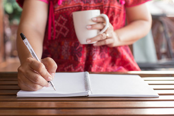 A woman holding a pen is writing on a notebook and a coffee mug on wooden table with warm light