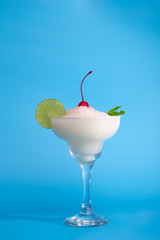 An isolated Margarita cocktail with a garnish lime, mint leaf and a cherry on top on a blue background