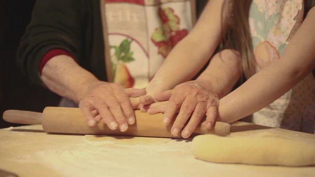 Elderly woman a young girl preparing pasta a cake or pizza together using a rolling pin on a sheet of dough.
