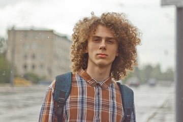 curly-haired young guy stands on the street with a satchel on his shoulder. student and young...