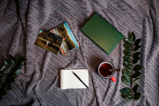 Red cup of tea, printed photos and old books on the gray plaid. Flat lay with old green books, pen, green leaves and printed photos.