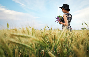 Cute attractive girl with a bouquet of colorful flowers in her hands. Young woman breathes in the scent of plants on wheat field during sunset. Pensive look. Romantic atmosphere.