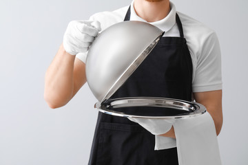 Handsome waiter with tray and cloche on light background