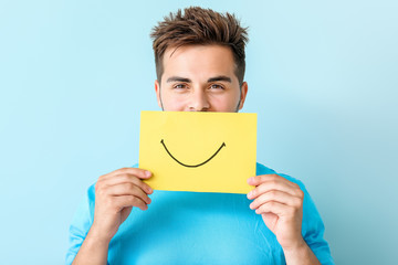 Happy young man with drawn smile on sheet of paper against color background