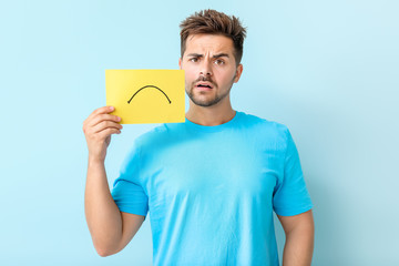 Sad young man with drawn upset mouth on sheet of paper against color background