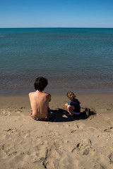 Young father and little daughter sitting on the beach and playing with sand.