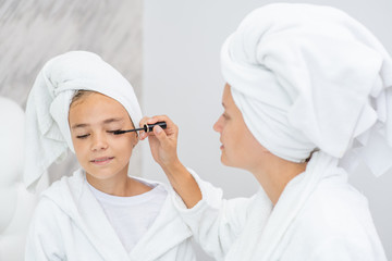 Happy family at home. Mother and young daughter while applying makeup at home. Mom and child girl are in bathrobes and with towels on their heads