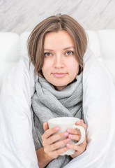 Beautiful sick woman sitting on the bed wrapped in a white blanket and holding a mug of hot tea