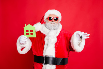 Portrait of cheerful santa claus in eyewear eyeglasses holding keys and paper card building give move house present wearing hat cap belt isolated over reed background