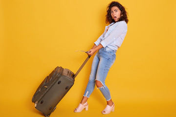 Attractive woman carries heavy suitcase, female with open mouth wearing casual shirt ana jeans, being late for plane, having shockes facial expression, standing isolated over yellow background.