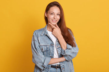 Close up portrait of Caucasian woman wearing stylish outfits stands smiling isolated over yellow background, female keeps hand under chin, having dark straight hair, looking directly at camera.