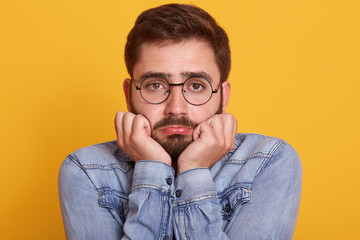 Unhappy offended young bearded male curves lower lip and keeps hand under chin, posing against...