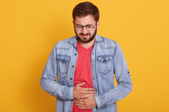 Close Up Studio Portrait Of Upset Sad Worried Guy Having Stomach Ache Disorder, Troubled Man Wearing Denim Jacket And Red Casual Tshirt, Posing Isolated Over Yellow Background. Health Care Concept.