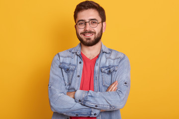 Close up portrait of bearded man with folded hands, sytanding and looking directly at camera with happy facial expression, being in good mood, handsome male dressed stylish clothing. People concept.