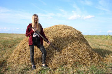 Naklejka na ściany i meble Joyful girl farmer in a burgundy sweater, in the working process, harvests hay in a field with a pitchfork in her hands.