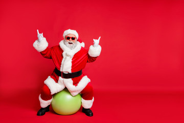 Full length body size view of his he nice attractive cheerful cheery glad fat bearded Santa working out sitting on pilates ball showing horn sign heavy metal isolated over bright vivid shine red