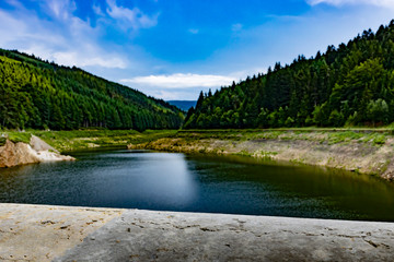 Image of an old concrete fence and lake with the beauty of the blue sky and the slopes covered by the green forest in the background.