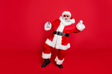 Forever young. Full length photo of excited santa in hat using cell phone device raising fists screaming yes have party wearing belt eyeglasses eyewear isolated over red background