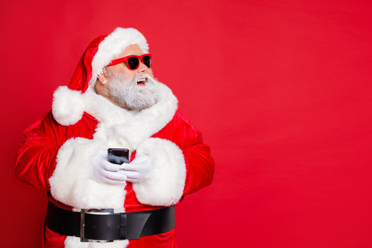 Portrait of cheerful santa claus in hat looking dreamy using cell phone plan newyear party wearing belt eyewear eyeglasses isolated over red background