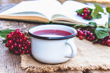 White mug or cup of hot viburnum tea on a wooden table near an open book and red viburnum berries. Source of natural vitamins. Used in folk medicine.