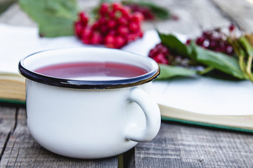 White mug or cup of hot viburnum tea on a wooden table near an open book and red viburnum berries. Source of natural vitamins. Used in folk medicine.
