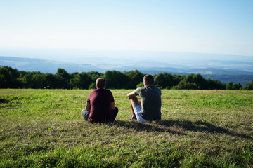 Fototapeta na wymiar Gay couple or two friends sitting on the grassy hill and watching romantic sunset
