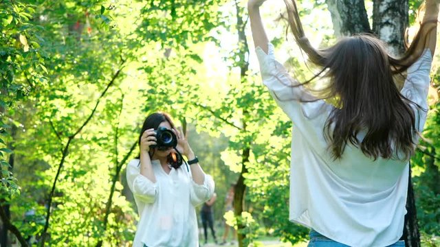 Pretty girl professional photographer wearing white shirt is making photos of a happy smiling girl in a park on a soft background of green foliage and spraying water.