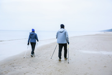 People jogging on the cold beach. Nordic walking with poles. Winter season at the seaside. Sporty couple. - obrazy, fototapety, plakaty