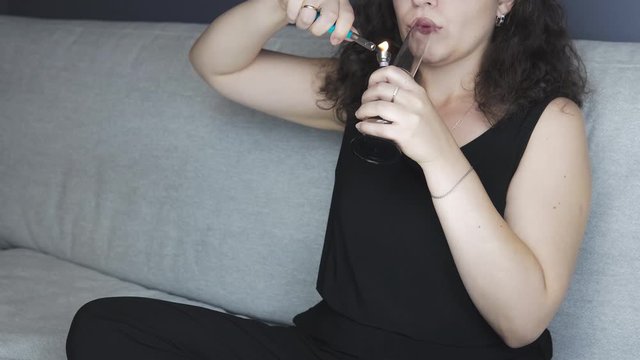The young person smoking medical marijuana with bong, indoors. The young woman smoke cannabis at home. Cannabis is a concept of herbal medicine.
